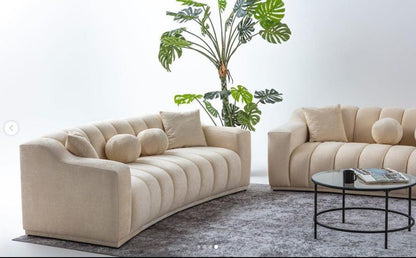Beżowa, tapicerowana sofa o nowoczesnym, falistym kształcie z pionowymi przeszyciami.