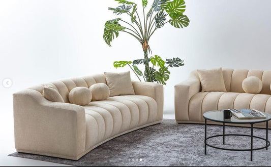Beżowa, tapicerowana sofa o nowoczesnym, falistym kształcie z pionowymi przeszyciami.